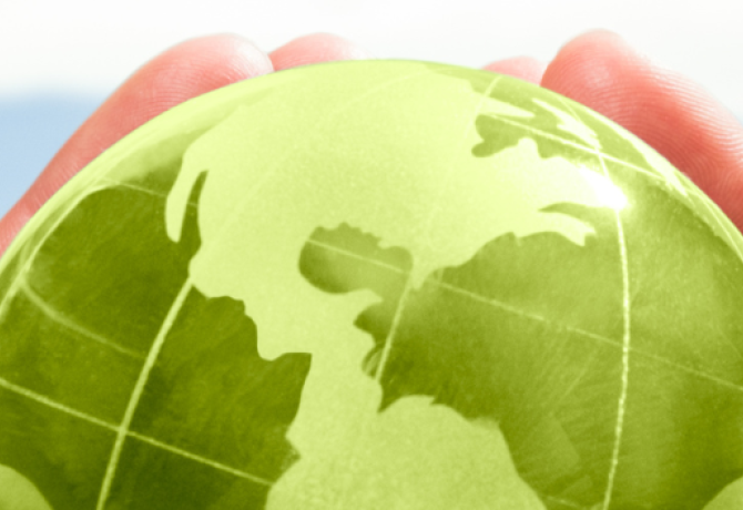 hands holding a small green globe in the foreground with rolling hills and grass in the background