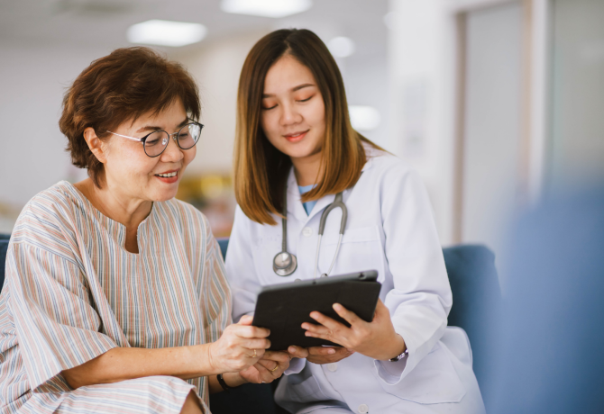 An elderly individual and a young health care professional sit on a couch looking at tablet they both hold in their hands.