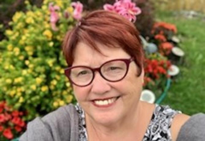 Dorothy, a woman with red hair and wearing glasses, smiles at the camera. There are flowers in the background.